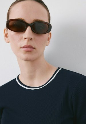 Ženska s svetlo poltjo in lasmi, zglajenimi nazaj, ki nosi črna ovalna sončna očala in mornarsko modro majico z rebrastim vzorcem in belim robom, na enobarvnem ozadju.