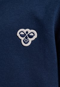 Mörkblå tröja i tyg med texturerat väv. Har en vit broderad bi-logga med blå detaljer på vänster sida.