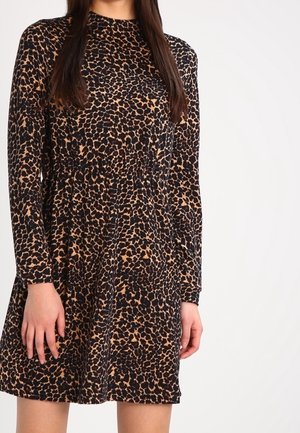 Femme portant une robe à manches longues, au-dessus du genou, imprimé léopard avec un col rond, debout devant un fond clair uni.