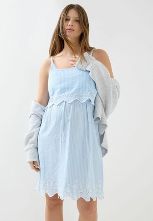 Žena v svetlomodrom bezrukávovom šate s bielymi kvetinovými výšivkami a sivým kardigánom prehodeným cez ramená.