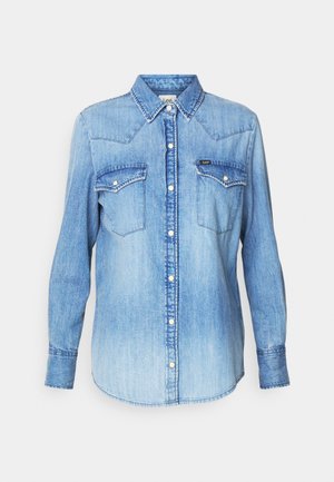Chemise en denim bleu clair à manches longues, col pointu, deux poches poitrine, fermeture à boutons et décoloration subtile du tissu. Étiquette de la marque visible.