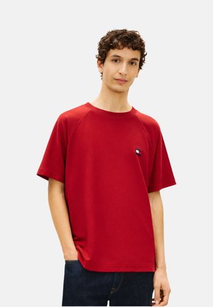 Joven con camiseta de manga corta roja con un pequeño logo en el pecho, posando con una mano en el bolsillo frente a un fondo liso.