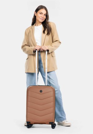 Kvinde i beige blazer og jeans, der står med hænderne på håndtaget af en brun kuffert på hjul mod hvid baggrund.