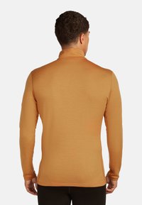 Mann von hinten gesehen, der einen taillierten langen kamelfarbenen Rollkragenpullover und schwarze Hosen vor einem schlichten weißen Hintergrund trägt.