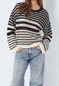 Gestreifter Strickpullover in Schwarz und Creme mit lockerem Design, weitem Schnitt und strukturierter Stoffqualität. Kombiniert mit hellblauen Jeans mit weitem Bein.