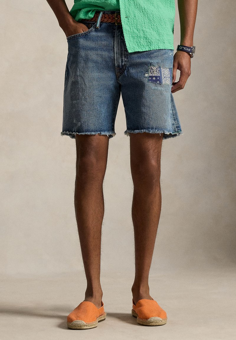 Moški, ki nosi obrabljene jeans hlače s trgačo modro obliž, zeleno majico, oranžne espadrile in črno zapestno uro z modrim paščkom.