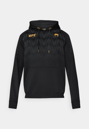 Must kapuutsiga dressipluus, millel on tekstuuriga muster, ees tasku, nööriga kapuuts ja kuldsed aktsendid. Rinnal UFC logo.