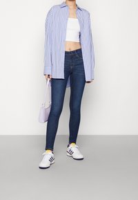 Light blue striped oversized shirt, white crop top, dark skinny jeans, purple-accented sneakers, and small lavender handbag.