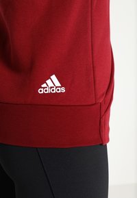 Gros plan de l'ourlet d'un sweat-shirt rouge Adidas sur des leggings noirs, montrant le logo blanc Adidas sur le tissu.