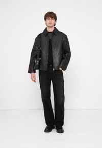 Veste en cuir noire avec col, fermeture éclair et poches latérales. Portée sur une chemise noire et associée à un jean sombre et des chaussures noires.