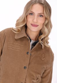 Ung kvinde med blond bølget hår iført en brun kordfløjljakke med ankerbroderi og en ternet skjorte under, som smiler blidt.