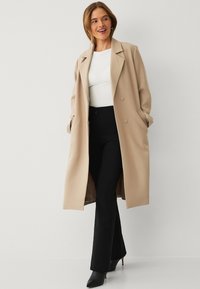 Beige dubbelzijdig jasje met grote revers, zijzakken en een gladde textuur, gedragen over een wit topje en zwarte broek, gecombineerd met zwarte hakken.