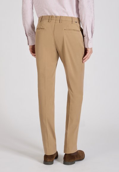 Uomo con pantaloni beige, scarpe di camoscio marroni e camicia rosa a righe a maniche lunghe infilata dentro, in piedi con le spalle rivolte verso l’osservatore su sfondo semplice.