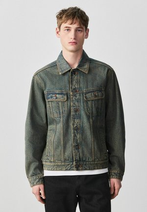 Giovane uomo con giacca di jeans blu slavata, con davanti bottoni e tasche sul petto, in piedi davanti a uno sfondo semplice grigio chiaro.