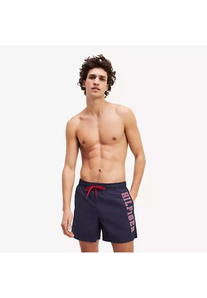 Oben ohne junger Mann, der marineblaue Badeshorts mit rotem Kordelzug und dem Schriftzug "HILFIGER" am linken Bein trägt, steht mit einer Hand in der Tasche.