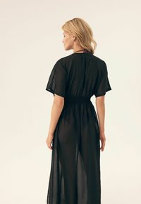 Czarna, przezroczysta, długa sukienka z krótkimi rękawami, z wcięciem w talii i zwiewną sylwetką. Posiada gładką fakturę i minimalne zdobienia.