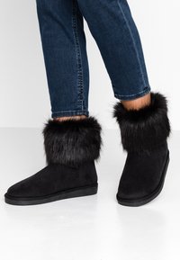 Stivaletti neri alla caviglia in suede con un caldo polsino in pelliccia sintetica nera. Suola piatta, punta arrotondata e senza hardware o motivi visibili.