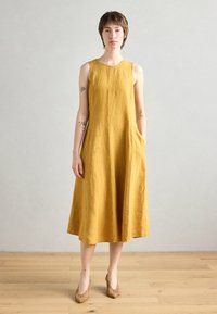 Gul leinen uten ermer kjole med avslappet passform, sidelommer og kne-lengde hem. Kledd med beige mønstrede hæler på et tregulv.