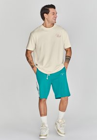Camiseta de manga corta color crema con un logotipo, combinada con pantalones cortos azul-verde con detalles laterales, cordón blanco y zapatillas beige.