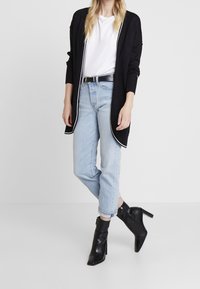 Černý cardigan s bílým lemem, bílé tričko, světle modré džíny a černé kotníkové boty na podpatku. Ležérní střih s rovnými nohavicemi.