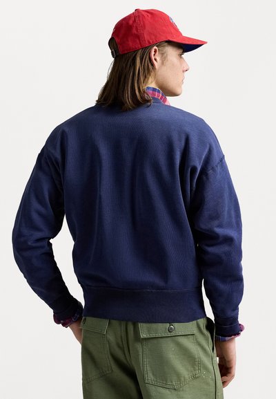 Sweat-shirt bleu marine avec l'ourlet et les poignets côtelés, en tissu texturé, associé à un pantalon vert. Casquette rouge avec logo et chemise à carreaux visible en dessous.