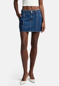 Mini-jupe en denim bleu foncé avec une taille ajustée, des poches avant et des accents de boutons argentés, présentant un design simple et épuré.