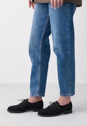Person trägt blaue Jeans aus Denim und schwarze Wildleder-Schnürschuhe und steht auf einem hellen Boden vor einem weißen Hintergrund.