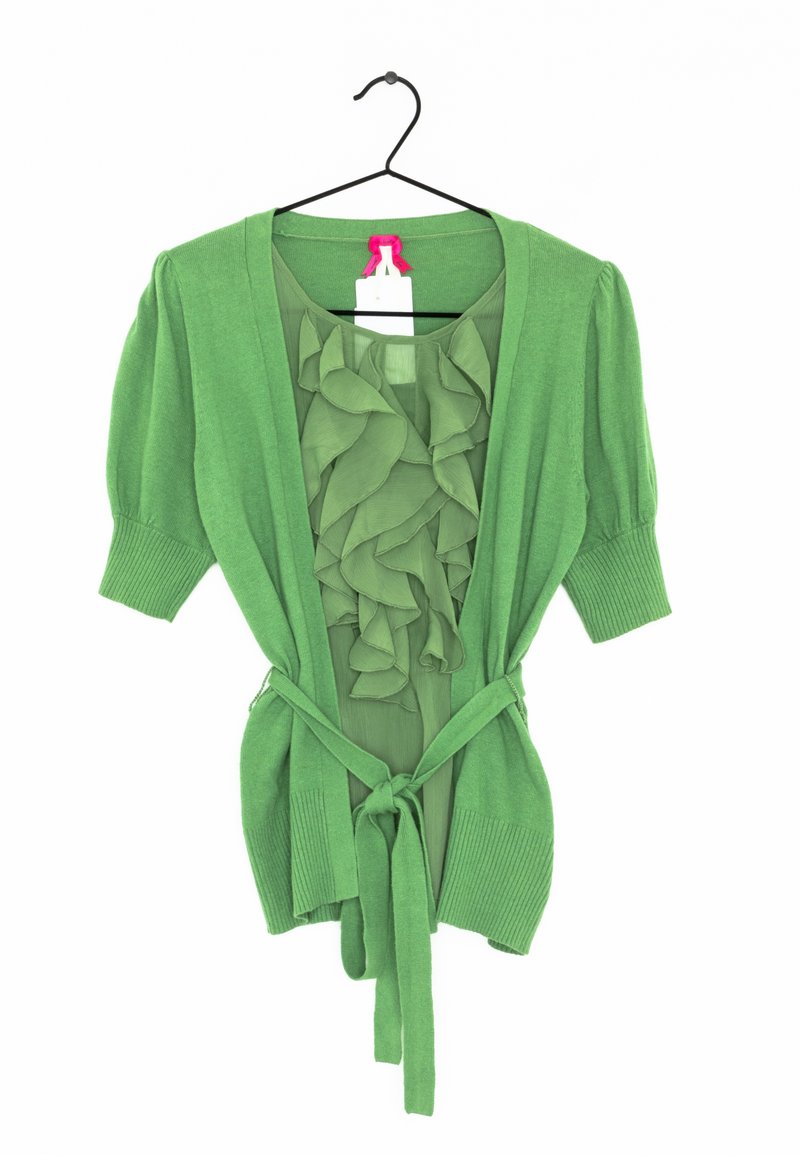 Cardigan vert à manches courtes avec une ceinture nouée à la taille, superposé à un chemisier vert à volants, suspendu sur un cintre noir.