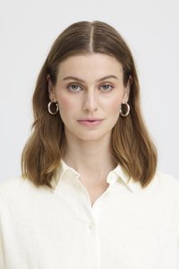 Žena s hnědými vlasy po ramena, která nosí stříbrné kruhové náušnice a bílou košili na knoflíky, čelí dopředu s neutrálním výrazem.