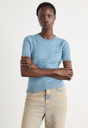 Lys blå ribbet strikkegenser med kabeldesign, korte ermer og rund hals, kombinert med lyse denimjeans.