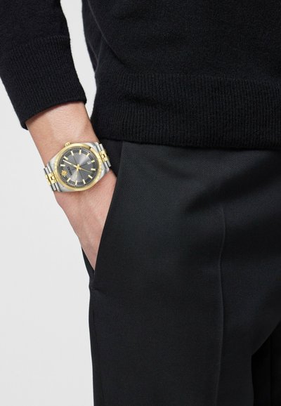 Poignet portant une montre bicolore argent et or avec un cadran sombre, à côté d'une manche de pull noir et d'une poche de pantalon noire avec la main à l'intérieur.