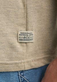 T-shirt en coton beige clair avec une étiquette rectangulaire cousue sur le côté, présentant un texte bleu et une couture bleue le long de l'ourlet.