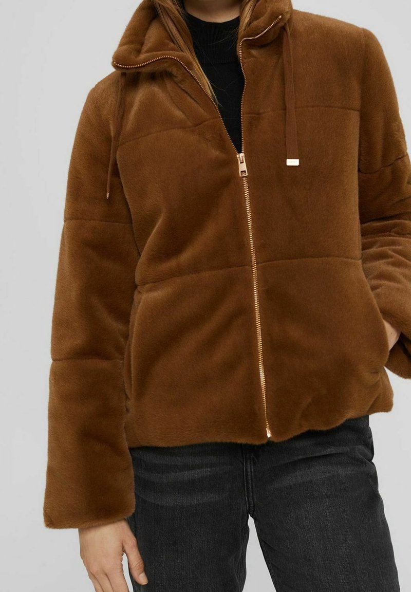 Veste marron duveteuse zippée avec col montant portée sur un haut noir et un jean foncé, main dans la poche, sur fond uni.