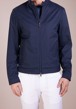 Homme portant une veste zippée bleu marine avec col montant et pantalon blanc, debout devant un fond uni.