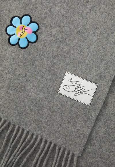 Grå uldbånd med en blå blomst-applikation med et smilende ansigt, og en hvid etiket med sort syning og en signatur. Frynser i kanten.