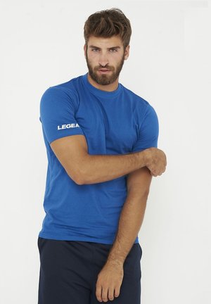 Mann mit Bart, der ein blaues LEGEA T-Shirt und dunkle Hose trägt, richtet seinen linken Ärmel vor weißem Hintergrund.