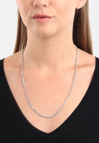 Kvinna som bär ett silverkedjehalsband över en svart V-ringad topp, med rakt brunt hår synligt bakom axlarna.