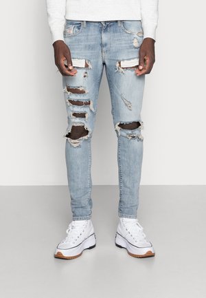 Personne portant un jean skinny bleu clair déchiré et des baskets blanches à plateforme, debout devant un fond clair uni.