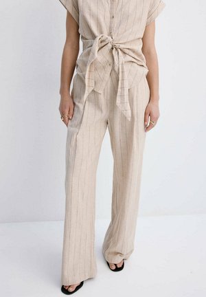 Frau trägt beige breit geschnittene Hosen mit Nadelstreifen und ein passendes kurzärmeliges Oberteil, das in der Taille gebunden ist, mit schwarzen offenen Sandalen.