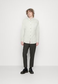 Camicia button-up verde chiaro, maniche lunghe, taschino frontale, abbinata a pantaloni grigio scuro e scarpe nere. Tessuto liscio, design semplice.