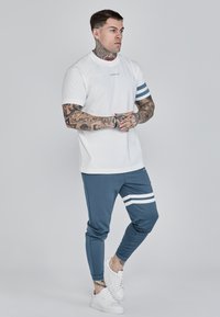Bílá tričko s modrými pruhy na rukávech, spárované s modrými tepláky se bílými bočními pruhy. Model má na sobě bílé tenisky.
