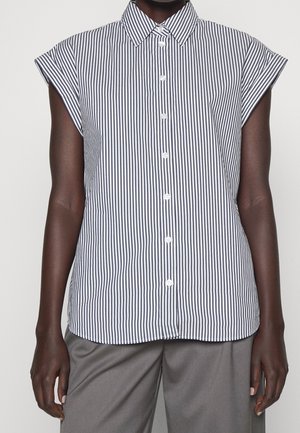 Femme portant une chemise à manches courtes rayée verticalement noir et blanc avec un col pointu et un pantalon gris.