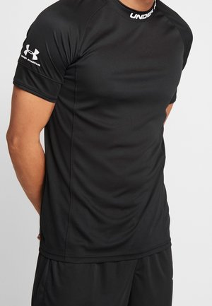 Homme portant un t-shirt de sport noir Under Armour à manches courtes avec un logo sur le col et la manche gauche, debout, les mains derrière le dos.