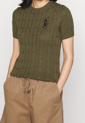 Personne portant un pull court olive à torsades, avec un petit logo brodé, et un pantalon beige à cordon.