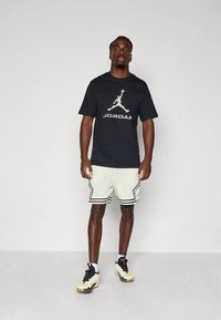 Svart bomulls-T-skjorte med hvit "JORDAN"-logo; lysegrønne mesh-shorts med svarte striper; svarte og gullfargede joggesko med hvite detaljer.