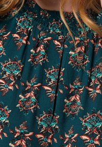 Vestido floral en color verde azulado que presenta un escote fruncido, con patrones florales en rojo y turquesa y un detalle de pliegue vertical en el frente.