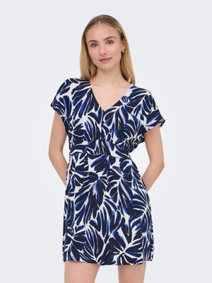 Jeune femme en robe à manches courtes et col en V avec des motifs abstraits de feuilles bleues et noires, debout, les mains dans les poches, sur un fond clair.
