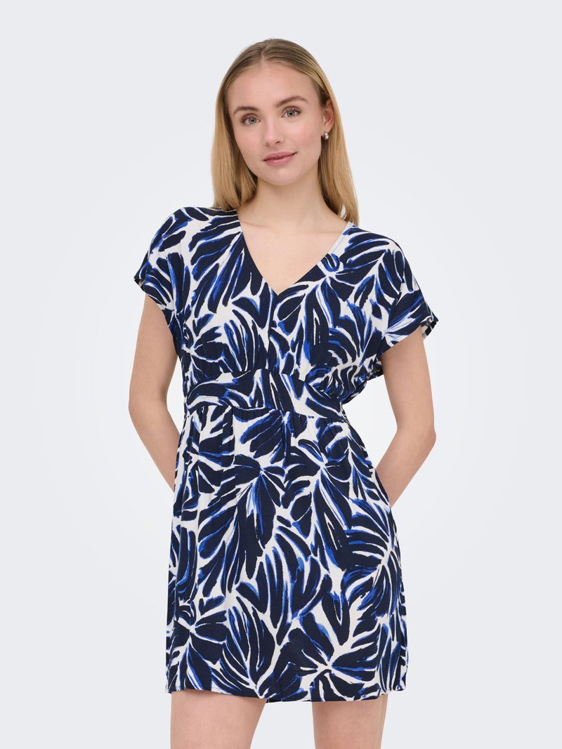 Jeune femme en robe à manches courtes et col en V avec des motifs abstraits de feuilles bleues et noires, debout, les mains dans les poches, sur un fond clair.