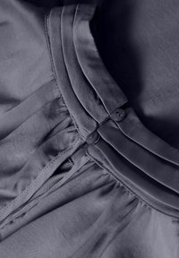 Gros plan d'un vêtement en tissu foncé avec col plié, deux petits boutons ronds et surpiqûres froncées près de la couture.