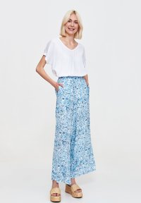 Biały krótki rękaw bluzka w połączeniu z niebieskimi szerokimi spodniami w kwiatowy wzór. Modelka nosi plecione sandały na platformie.